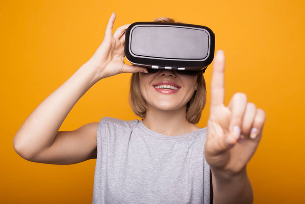 Front view photo of a blonde woman with vr eyeglasses touching something on a yellow studio wall and smile - Photo, Image