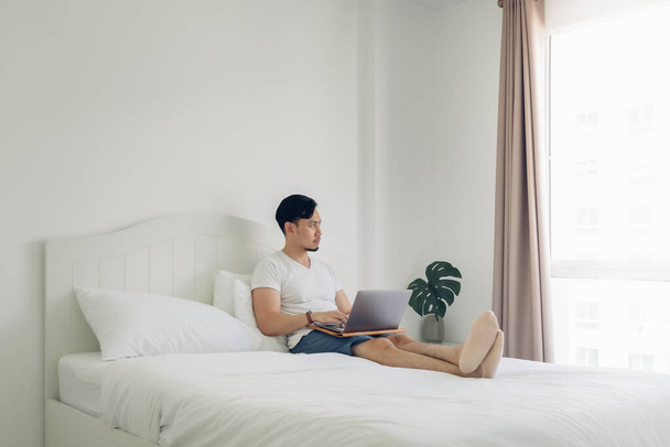 Man lying on bed work on his laptop in cozy white bedroom. - 写真・画像