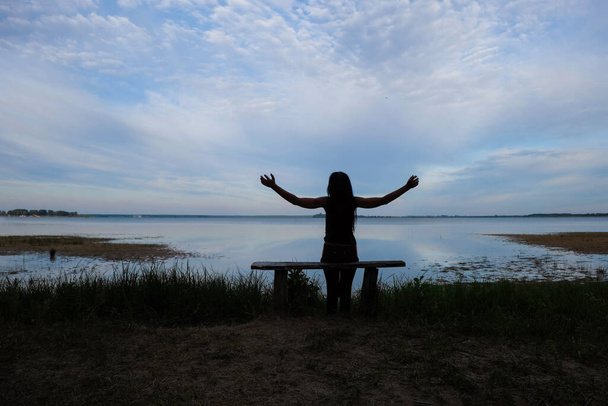 Силуэт женщины с вытянутыми руками. Женщина, сидящая на скамейке на фоне озера. Рассвет на украинском озере Свитязь. Копирование пространства. - Фото, изображение