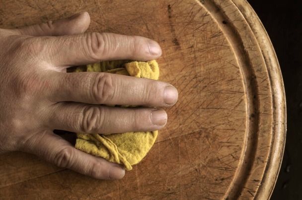 Зріла чоловіча рука протирає ріжучу дошку кухонною ганчіркою. Крупним планом чоловіча рука з жовтою ганчіркою. Гендерний стереотип. Вибірковий фокус. Тонована фотографія
 - Фото, зображення