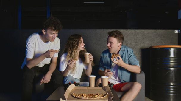 Молодые друзья едят пиццу и разговаривают в темной комнате - Фото, изображение