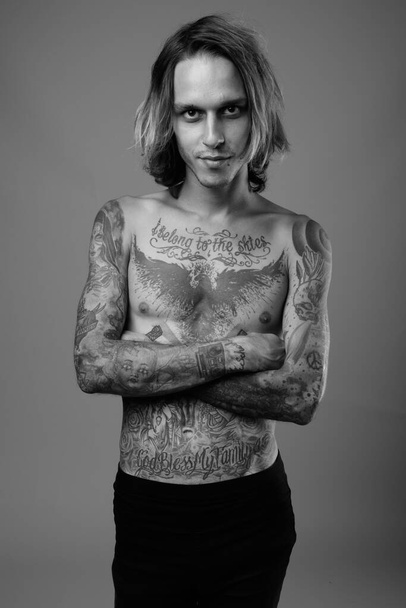 Studio ujęcie zbuntowanego młodzieńca bez koszuli z tatuażami na ciele na szarym tle w czerni i bieli - Zdjęcie, obraz
