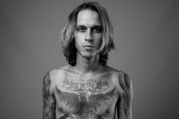 Estudio plano de joven rebelde sin camisa con tatuajes corporales sobre fondo gris en blanco y negro - Foto, imagen