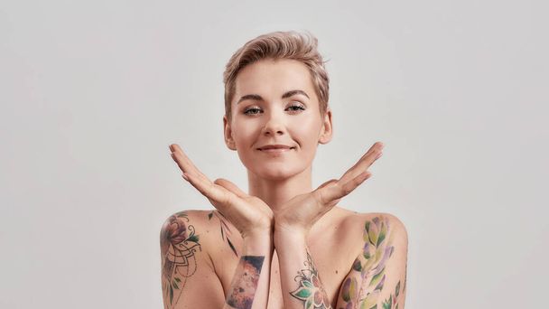 Portrait of half naked tattooed woman with short hair looking at camera, holding open palms under her chin isolated over light background - Фото, изображение