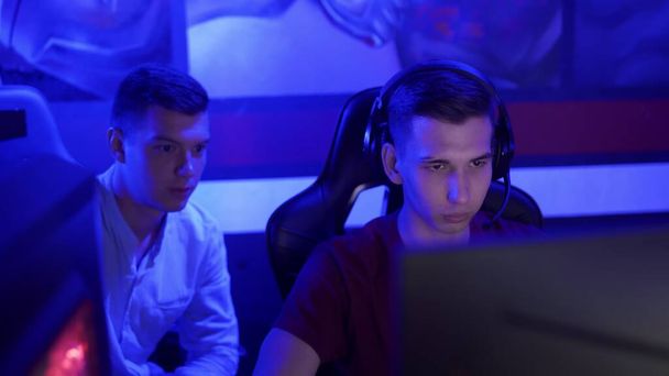 Mladý muž dává rady svému příteli, zatímco on hraje kolo videohry v moderním kybersportu klubu - Fotografie, Obrázek
