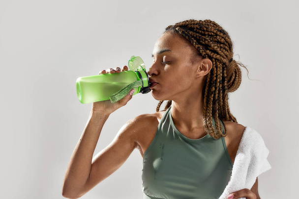 Drink it. Close up portrait of young sportive mixed race woman holding bottle, drinking protein shake cocktail after intensive training isolated over grey background - 写真・画像