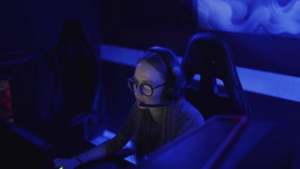 Skoncentrowana młoda kobieta w okularach i słuchawkach siedzi przed monitorem i gra w gry wideo w klubie komputerowym - Zdjęcie, obraz
