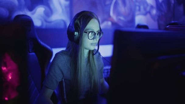 Geconcentreerde jonge vrouw met bril en koptelefoon zit voor de monitor en speelt in video game in computer club - Foto, afbeelding