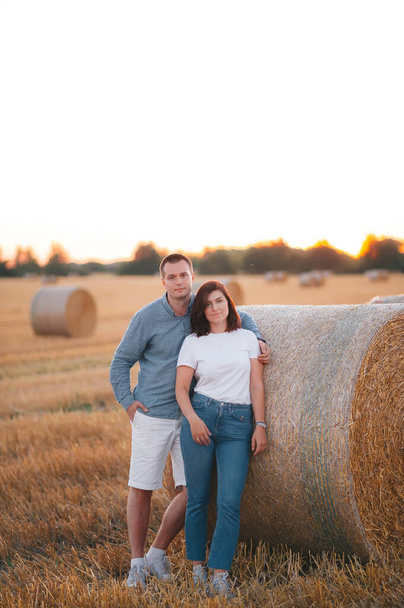 Das schöne Paar posiert nach der Ernte auf dem Feld und steht bei Sonnenuntergang in der Nähe eines großen Heuhaufens. Vertikales Porträt - Foto, Bild