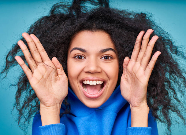 A close-up, cheerful, squeoring girl, with a large black curly hairdo, holds her hands near her face and smiles happily, looks affably at the camera, posing on a blue background. - Foto, Imagen