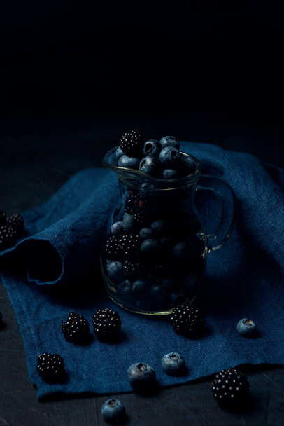 Dzbanek szklany z jagodami i jeżynami na niebieskiej serwetce i ciemnym, teksturowanym tle.  - Zdjęcie, obraz