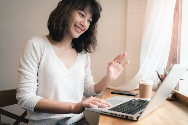 Young Asian woman working and video conferencing with laptop computer. A happy woman with smiley face working from home. A cup of coffee on table. Work from home concept. Bangkok, Thailand - 写真・画像