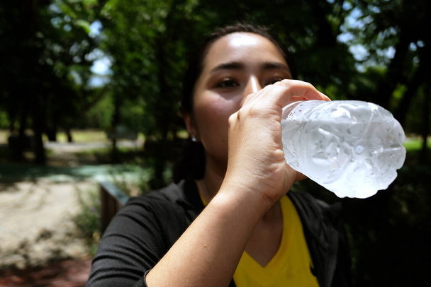 Asiatische junge Frau in schwarz-gelber Sportbekleidung Durstig und ausgeruht und trinkt nach dem Sport und dem Laufen im Garten Wasser in der Flasche. Geschlossen und konzentriert zur Hand - Foto, Bild