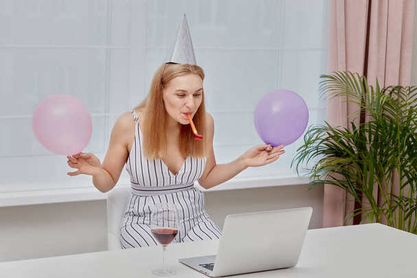 Happy blonde girl with glasses of wine celebrates a birthday in front of laptop. - Zdjęcie, obraz