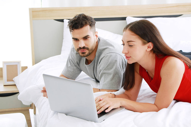 Gülümseyen Kafkasyalı çift, rahat beyaz yatakta sosyal medya teknolojilerinde internetten iş yapıyor ve dinleniyor. Evden çalışmak ve tatilde yaşam tarzı kavramı. - Fotoğraf, Görsel