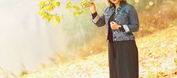 湖の近くの秋の公園で白人の女の子が身に着けている木の近くに立っています:青いデニムジャケットと黒ポルカドットドレス屋外で秋.   - 写真・画像