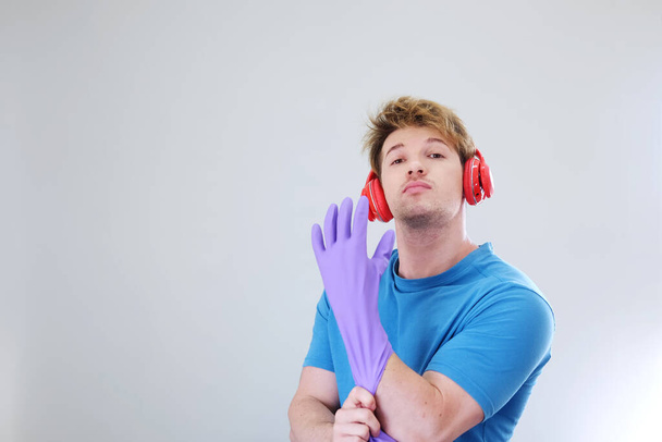 Улыбающийся ленивый молодой кавказский мужчина в фиолетовых резиновых перчатках для уборки и отдыха, слушая музыку в красных наушниках. Образ жизни семьи в праздничной концепции. - Фото, изображение
