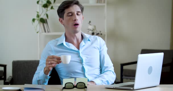 Jovem empresário freelancer sentado à mesa no escritório em casa, trabalhando no laptop, sentindo-se cansado, esticando, bocejando, bebendo xícara de café para animar, tentando se concentrar, conceito matutino - Filmagem, Vídeo