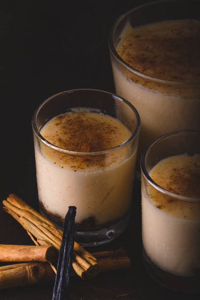 Three glasses of vanilla custard on wooden table, decorated with cinnamon stick and a strand of vanilla. Low key photography with matte finishes. - Photo, Image