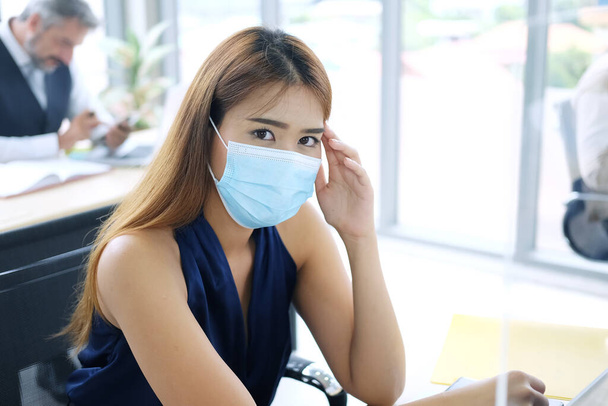 Молодая азиатская бизнесвумен в маске предотвращает распространение вируса Ковид-19, страдает головной болью и чувствует тошноту в офисе. Социальная дистанция и новая концепция нормального образа жизни - Фото, изображение