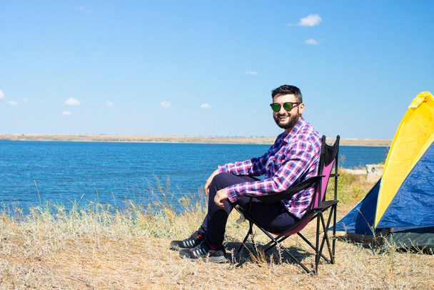 Cheerful handsome young bearded man tourist in sunglasses sitting in chair near touristic camping tent by the blue sea. Enjoying active summer or autumn vacation. Adventure travel lifestyle concept.  - Fotoğraf, Görsel