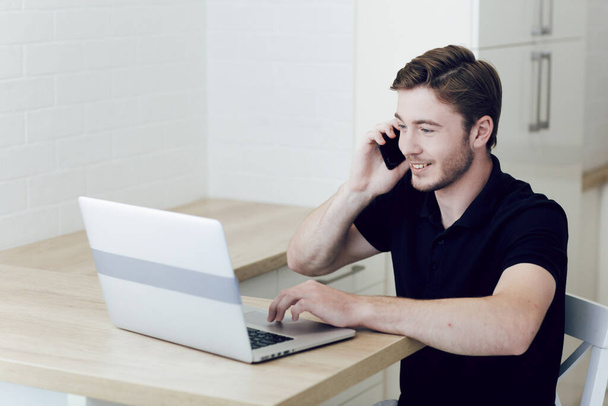 Молодой человек, работающий дома на ноутбуке, разговаривающий по мобильному телефону, сидящий за столом. Работа на расстоянии во время карантина коронавируса. Оставайтесь дома, самоизоляция - Фото, изображение