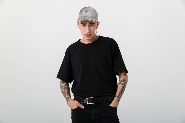 Portrait of bold young man wearing black clothes posing on camera with hands in pockets over white background - Foto, afbeelding