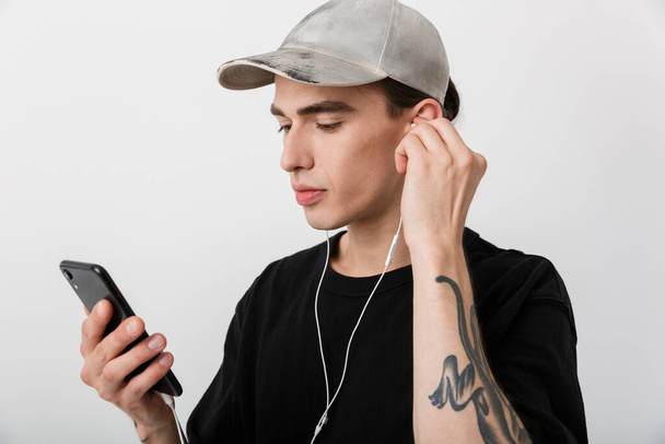Portret van een knappe man in zwarte kleren die naar muziek luistert met een mobiele telefoon en oortjes over een witte achtergrond - Foto, afbeelding