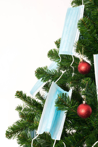 Imagen en color vertical con vista frontal de un árbol de Navidad decorado con bolas rojas y máscaras de coronavirus azul. Covid y feliz año nuevo concepto. - Foto, imagen
