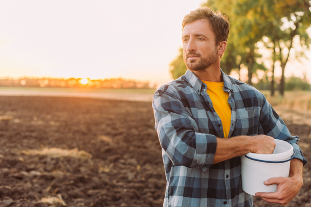 rančer v kostkované košili drží vědro, zatímco stojí na oraném poli ve slunečním svitu - Fotografie, Obrázek