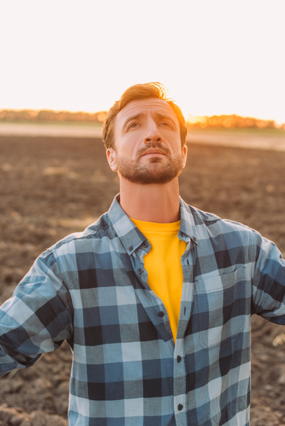 rancher in checkered shirt looking up while standing on plowed field - Fotoğraf, Görsel