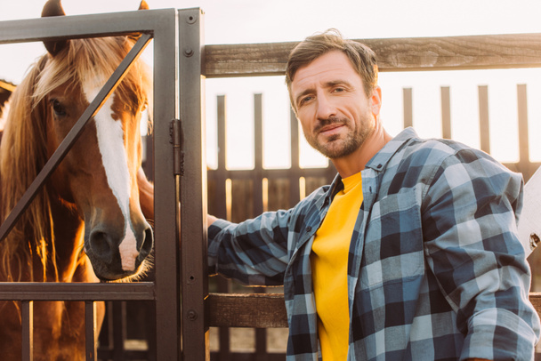 ranczer w koszuli w kratkę dotykając konia w zagrodzie i patrząc w kamerę - Zdjęcie, obraz