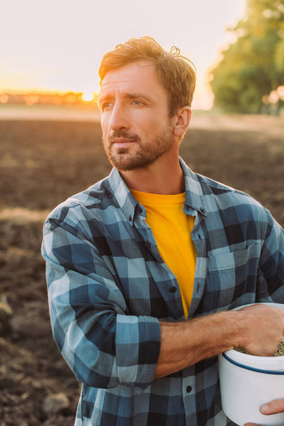 Ранчер в клетчатой рубашке смотрит в сторону, стоя на поле с ведром - Фото, изображение