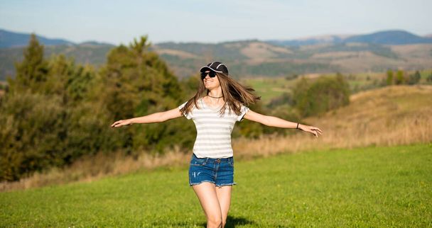 Piękna beztroska kobieta w okularach przeciwsłonecznych, czapce i dżinsach spaceruje po letnim polu z górami na tle - Zdjęcie, obraz