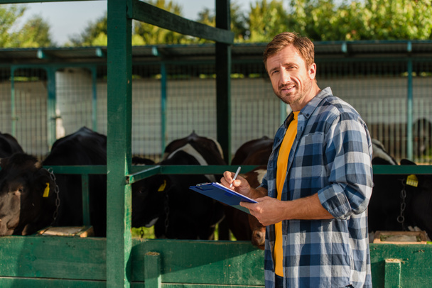 granjero en camisa a cuadros mirando a la cámara mientras está de pie cerca del establo y escribiendo en el portapapeles - Foto, Imagen