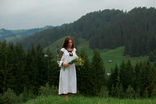 girl in embroidery with a bouquet of wild flowers stands on green grass in the carpathian mountains - Photo, Image