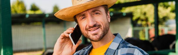 concepto horizontal de ranchero en sombrero de paja mirando a la cámara mientras habla en el teléfono inteligente  - Foto, imagen