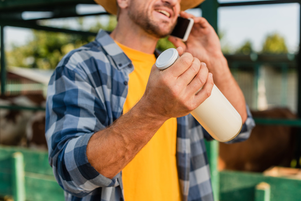μερική άποψη του γεωργού με καρό πουκάμισο μιλώντας στο κινητό τηλέφωνο και κρατώντας μπουκάλι φρέσκο γάλα, επιλεκτική εστίαση - Φωτογραφία, εικόνα