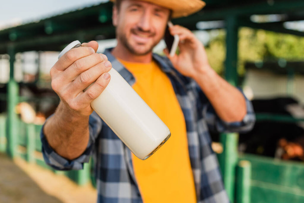επιλεκτική εστίαση του γεωργού μιλώντας στο smartphone και κρατώντας μπουκάλι φρέσκο γάλα, ενώ κοιτάζοντας κάμερα - Φωτογραφία, εικόνα