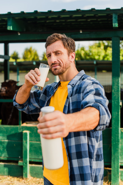 selektivní zaměření farmáře v kostkované košili drží sklo a láhev mléka při pohledu do kamery - Fotografie, Obrázek