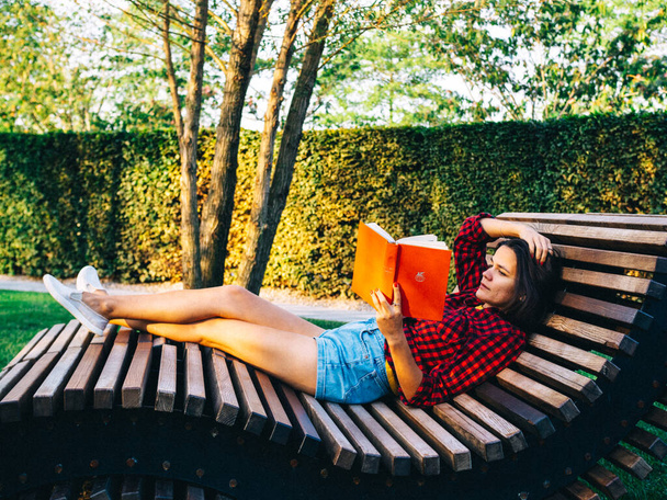 Joven chica caucásica se encuentra en el banco de la figura en el parque por la mañana y lee libro rojo - Foto, imagen