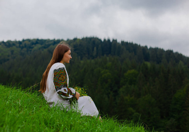 girl with loose hair in an embroidery with a bouquet of wild flowers sits on green grass against the background of the carpathian mountains - Φωτογραφία, εικόνα