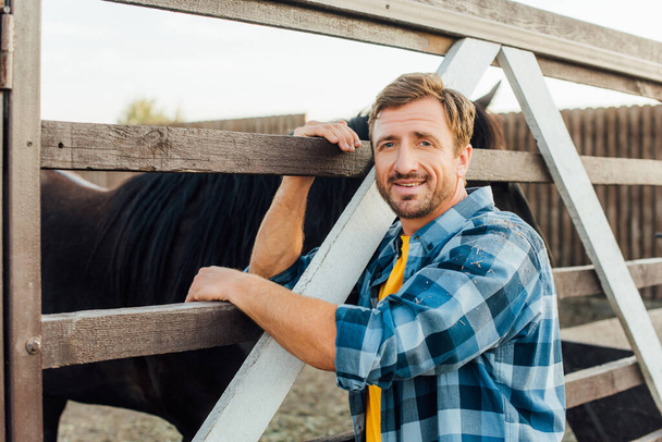 granjero con camisa a cuadros mirando a la cámara mientras se apoya en la valla de corral cerca de caballo - Foto, Imagen