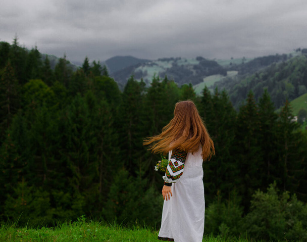 Девушка с распущенными волосами в вышивке с букетом полевых цветов счастливо бежит по зелени в карпатских горах - Фото, изображение