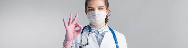A young beautiful woman doctor in a white coat, in a medical mask and pink sterile gloves shows OK sign on gray background. Concept of healthy man or trusting a doctor - Фото, зображення