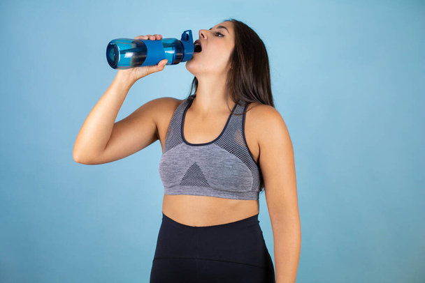 Молода красива спортивна жінка в спортивному вбранні посміхається щасливою і природною, п'є прісну воду з пляшки
 - Фото, зображення