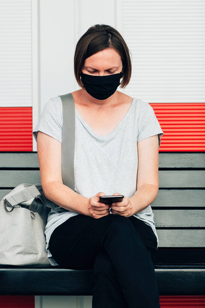 Mujer con mascarilla protectora escribiendo mensaje de texto en el teléfono inteligente mientras está sentado en el banco de la calle de la ciudad en el entorno urbano moderno - Foto, Imagen