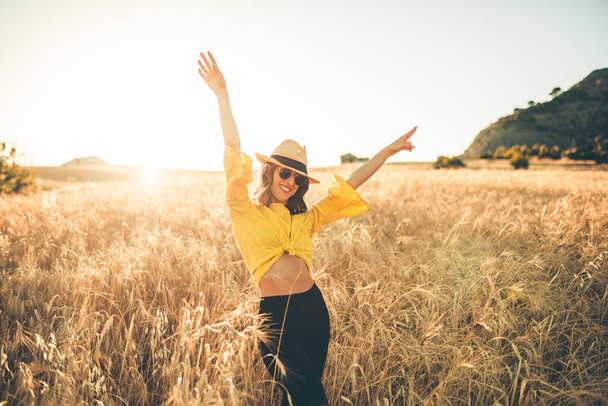 Молодая девушка, одетая в желтую соломенную шляпу, счастливо стоит посреди пшеничного поля на закате. - Фото, изображение