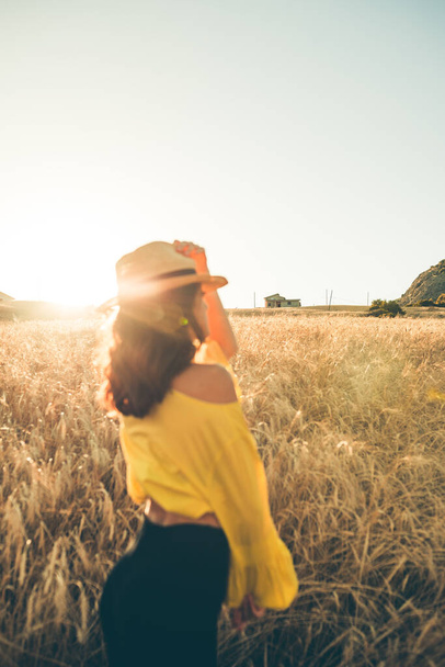 Chica joven vestida de amarillo con sombrero de paja se encuentra feliz en el medio del campo de trigo al atardecer. - Foto, Imagen