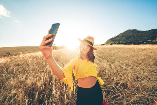 Junges Mädchen in Gelb mit Strohhut steht glücklich mitten im Weizenfeld bei Sonnenuntergang. - Foto, Bild
